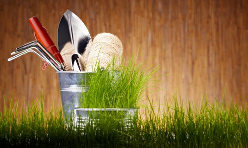 A close-up image of gardening tools and materials placed in a silver metal bucket set on a well-maintained grassy outdoor area, with vibrant green grass in the foreground and a warm wooden background. The tools include a hand rake, trowel, and cultivator, along with a ball of natural twine, indicating preparation or ongoing gardening activity. The scene suggests a neatly kept garden space, typical of residential properties in Bethnal Green, with natural lighting highlighting the fresh green foliage and the textures of the tools and grass, supporting professional gardening and landscaping services for maintaining vibrant and orderly outdoor gardens in east London.