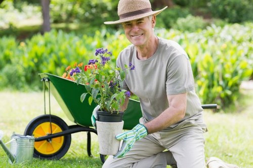 A smiling elderly man wearing a light grey t-shirt, beige trousers, a straw hat, and gardening gloves crouches on a well-maintained lawn in a residential garden. He holds a potted flowering plant with purple and red blooms, positioned in front of him. Behind him, a bright green wheelbarrow with a yellow wheel contains a few gardening tools and a metal watering can, suggesting outdoor maintenance work. The garden features a neatly trimmed hedge, lush green grass, and a backdrop of trees, providing a natural, peaceful environment. The scene is bathed in soft natural light, indicating a clear, pleasant day. This outdoor space emphasizes careful lawn care and plant nurturing, aligning with gardening services offered by Garden Maintenance Bethnal Green in the local Bethnal Green area of London, with the local postcode E2. The overall composition presents a tidy, vibrant garden poised for ongoing maintenance and horticultural care.