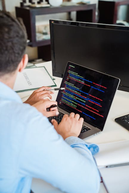A person working at a desk with a laptop displaying colorful lines of code, surrounded by a workspace including a monitor, a closed notebook, and a pen. The individual is sitting with their back parti